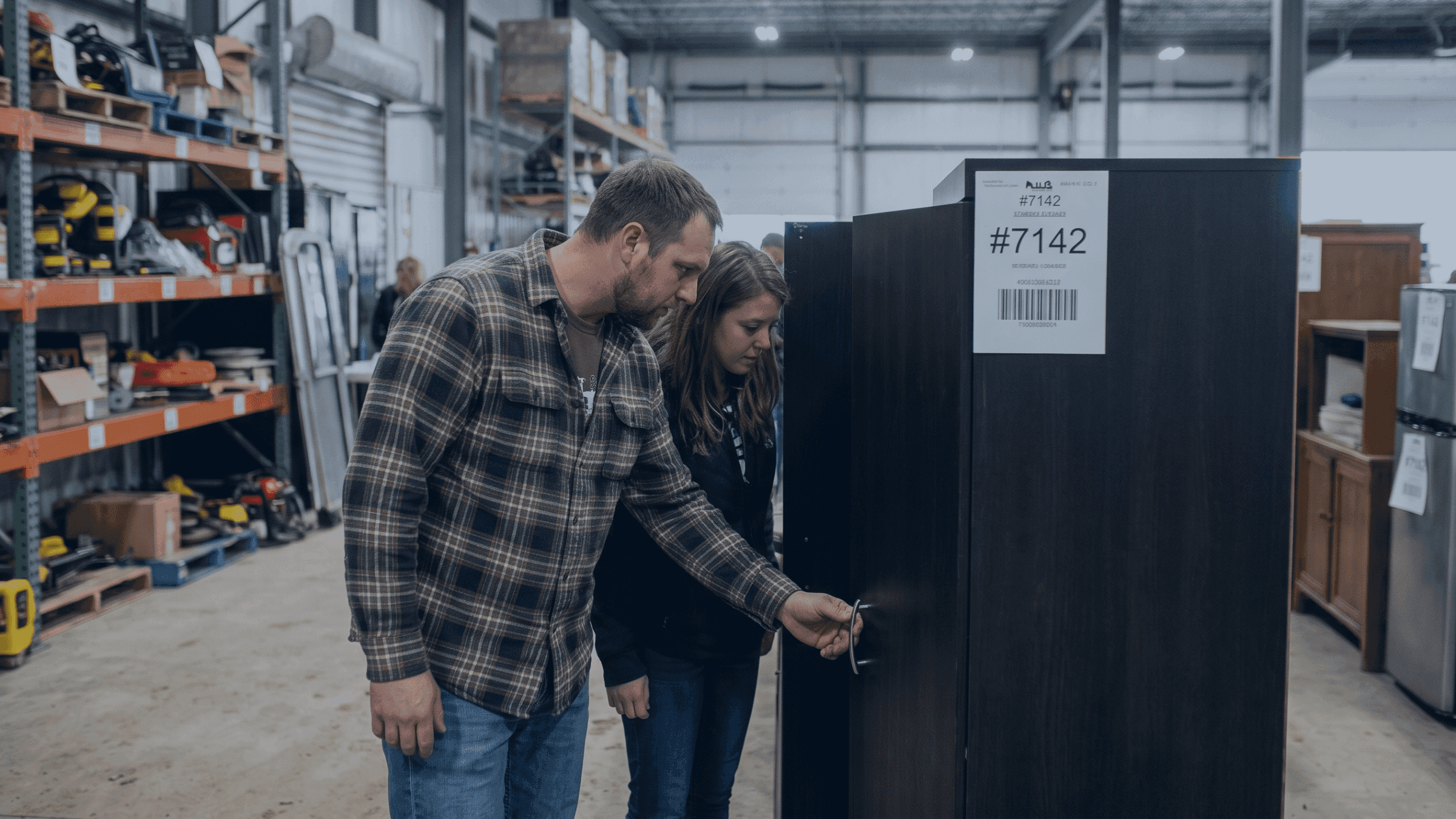 A man and woman inspect a dark cabinet, the man opens its door in a warehouse.
